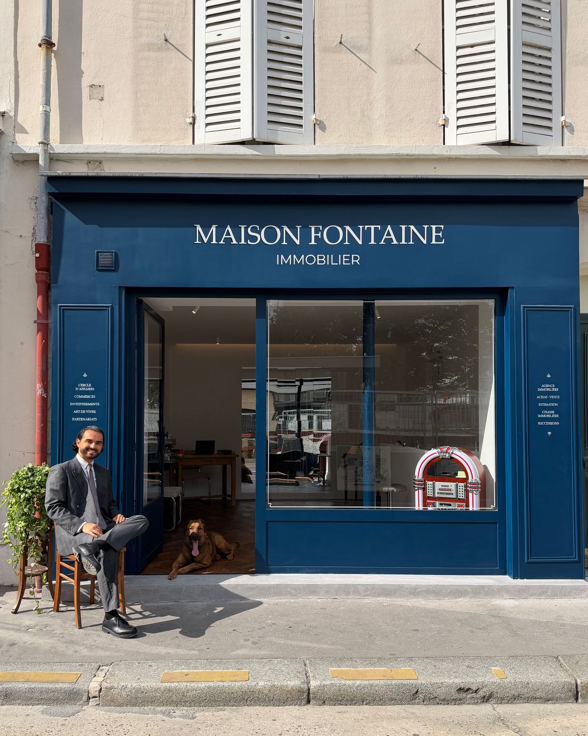 Fabien Fontaine, souriant, assis devant la devanture bleu marine de l'agence Maison Fontaine Immobilier, Thales couché sur le seuil